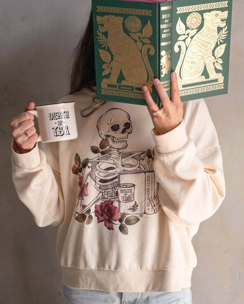 Model holding up a book with one hand a a WERKSHOP "Death by TBR" mug in the other hand. She is wearing a WERKSHOP "Death by TBR" hoodie in bone color. 