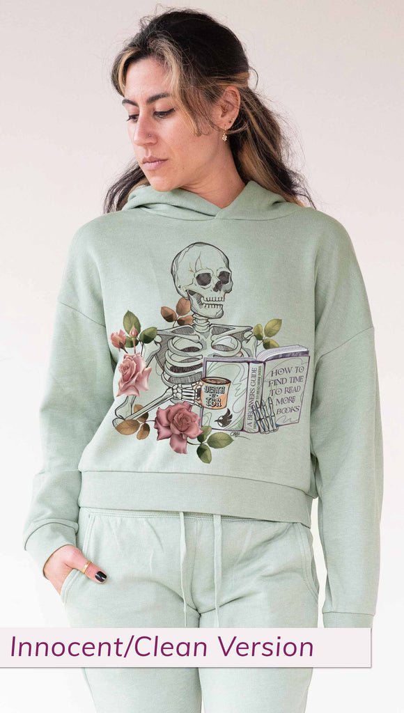 Model wearing WERKSHOP  "Death by TBR" Hoodie in Sage Colorway ~ Innocent/Clean version with the phrase, "How to Find Time to Read More Books". Featuring a skeleton holding a book in one hand and a coffee mug in the other with accent roses.